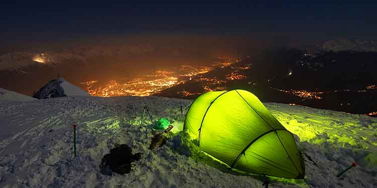 A four-season tent set up at a camp above the tree line in the snow overlooking a beautiful view of a city lit up at night.