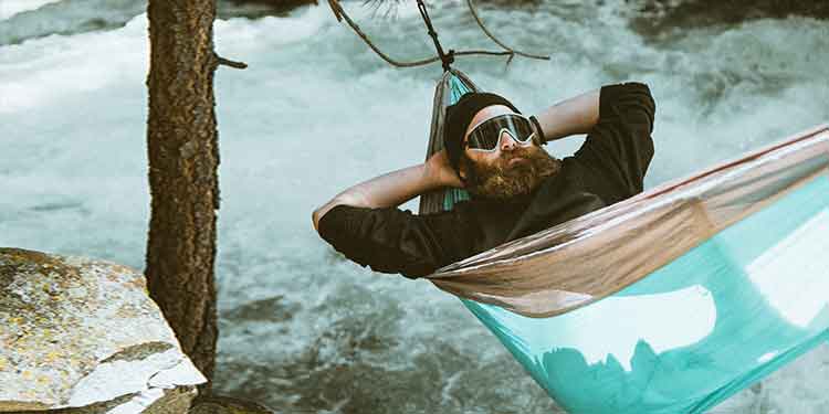 Bearded man relaxing on an outdoor adventure in a hammock by a river.
