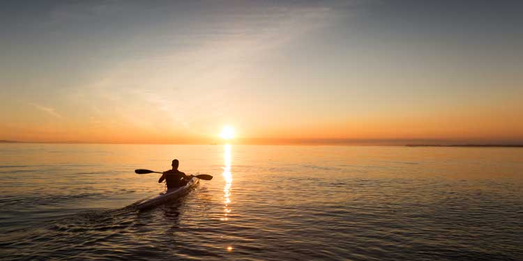 A man in a touring kayak with low-angle paddling style kayaking on still ocean water into the sunset.