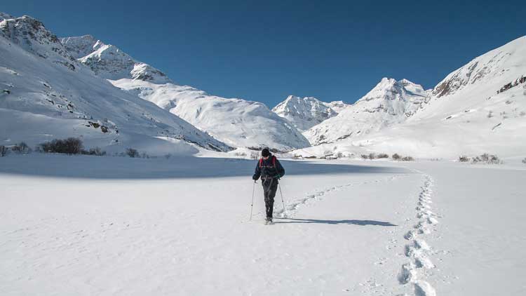 Man practicing proper snowshoeing safety techniques as he snowshoes in a snowy valley at the base of a mountain chain.