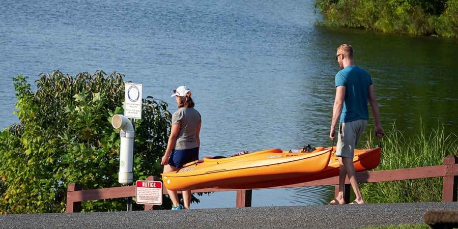 Two people carry two kayaks at the same time by use of the kayak grab handles.