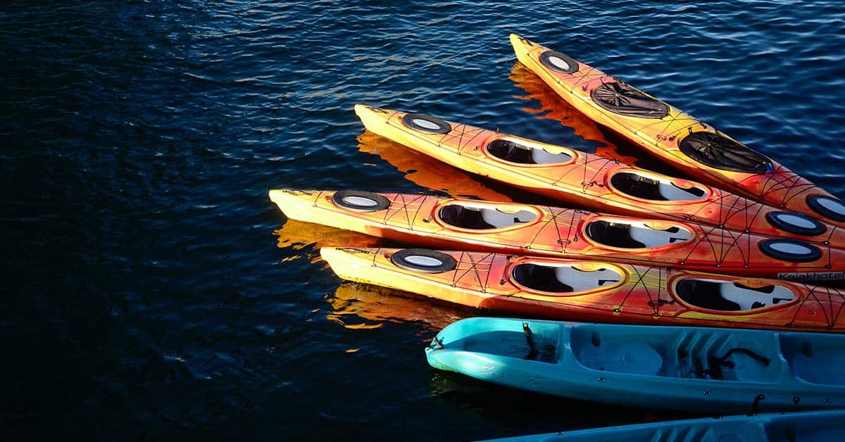 A group of sit-in 2-person kayaks and sit-on-top 2-person kayaks docked at a lake. Which of these tandem kayaks would be easier to paddle alone?