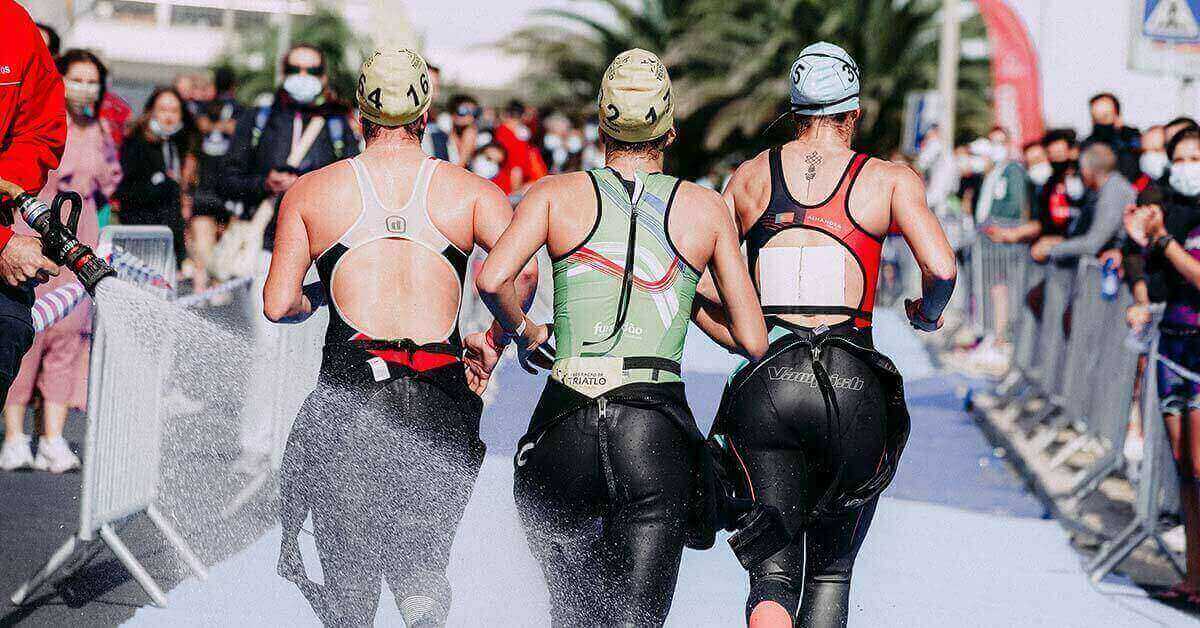Women running in a triathlon wearing one-piece tri-suits and swimsuits under their wetsuits.