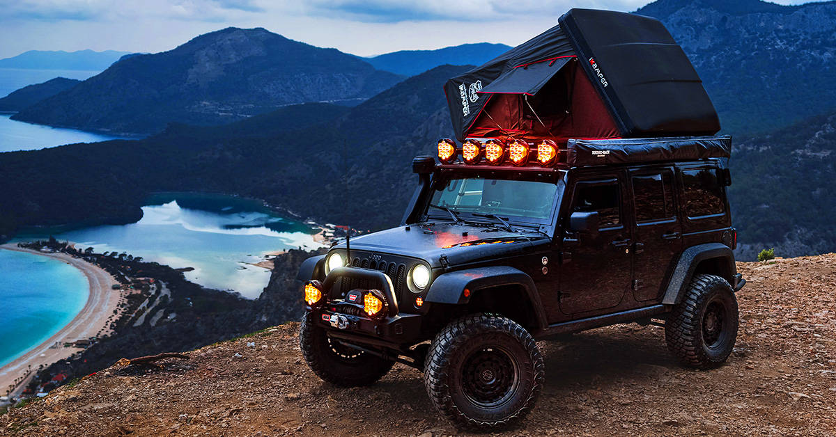 An iKamper hard shell tent set up on top of a 4-door Jeep Wangler parked on top of a ridge overlooking a beautiful view of the mountains and sea.