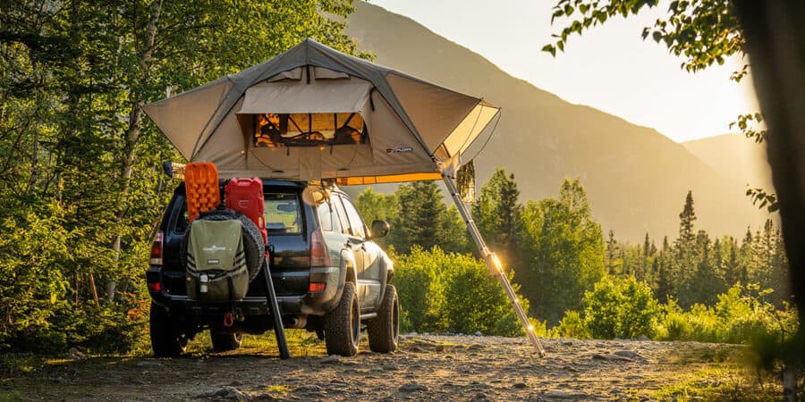 A soft shell roof-top tent on top of a 4x4 SUV in a wooded setting at sunset.