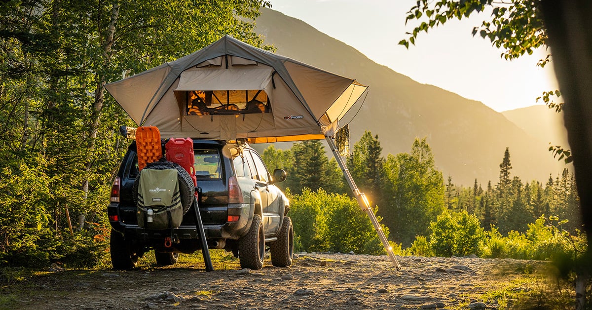 A soft shell roof-top tent on top of a 4x4 SUV in a wooded setting at sunset.