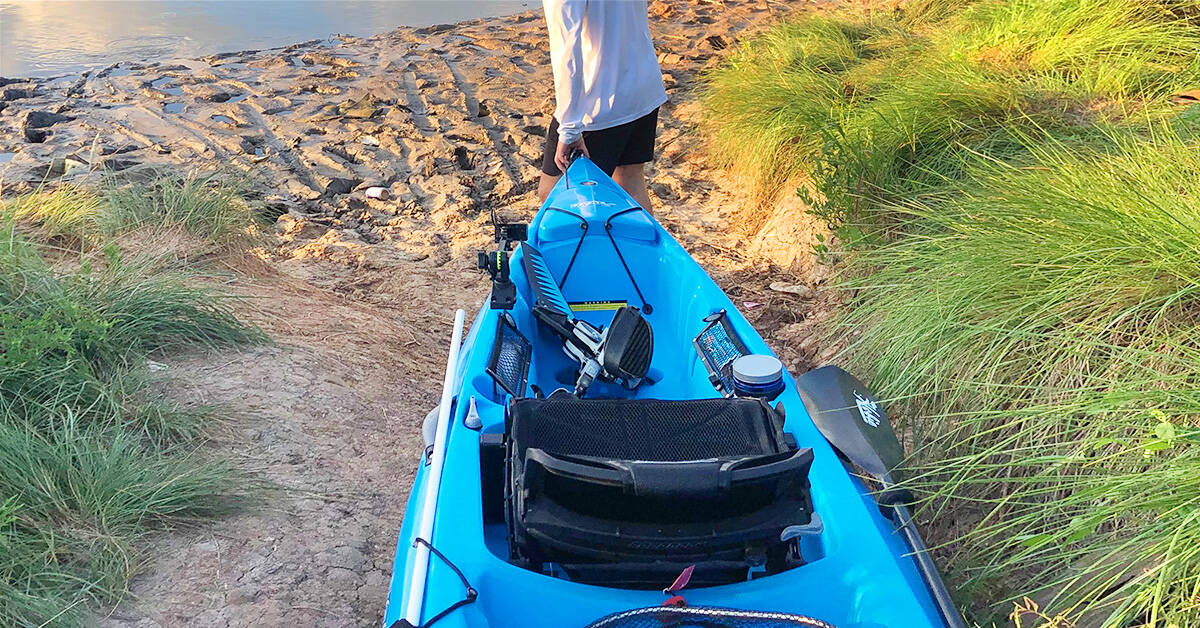 A Hobie pedal drive kayak loaded with fishing gear and a paddle being walked to the put in.
