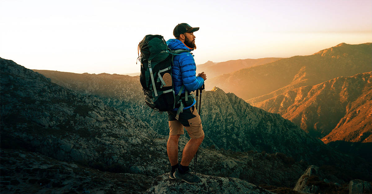 A backpacker with trekking poles on a hike at sunset in a mountain range.