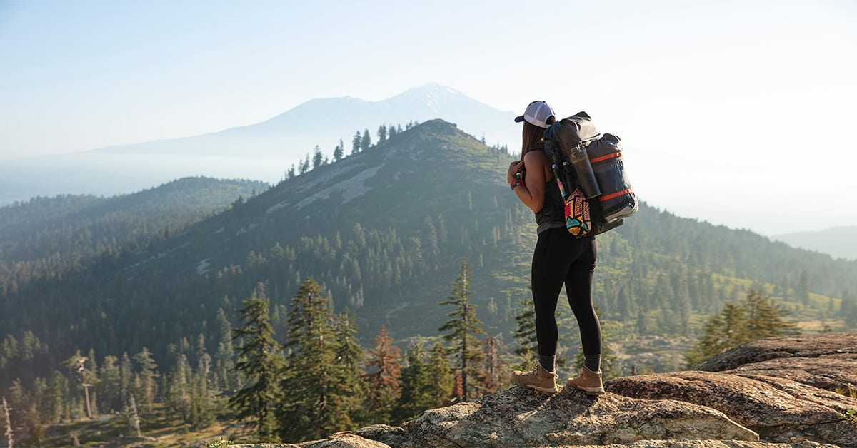 A female hiker wearing a heavy backpack and hiking boots looks over a scenic view in the mountians. 