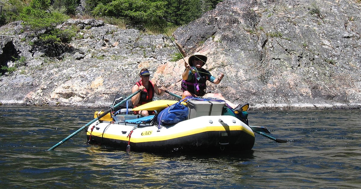 An NRS gear raft on a river with one man rowing and another one fishing from the front of the raft.