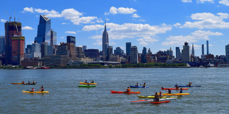 Kayakers paddle together in a large event on the Hudson River with New York City as the backdrop.