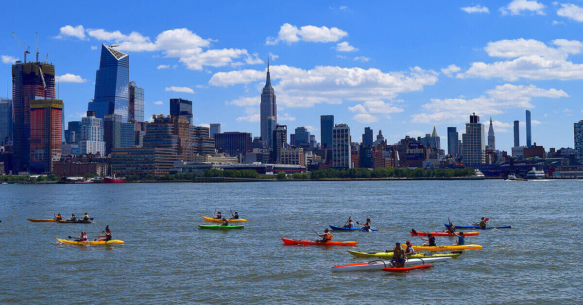 Kayakers paddle together in a large event on the Hudson River with New York City as the backdrop.