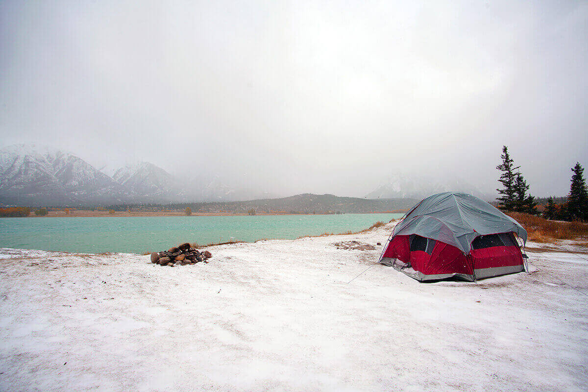 A red tent set up on the shore of a lake on a snowy winter day with mountains in the distance.
