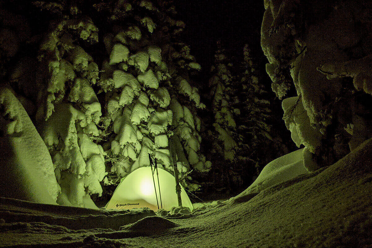 Skis and poles propped up by a tent in the woods at night after a heavy snowfall.