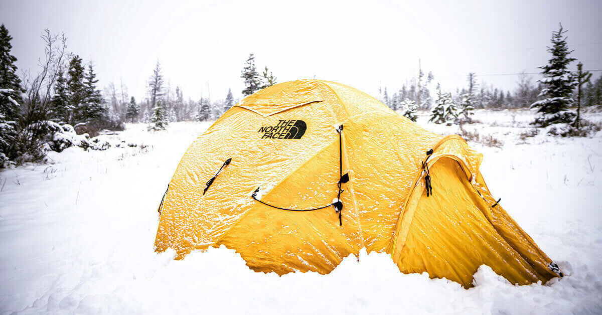 A a snowy wooded scene with a yellow, 4-season, insulated tent closed up for warmth.
