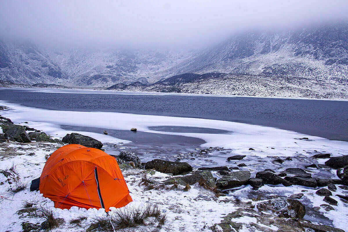 A orange tent set up in winter next to an icy lake at the foot of a mountain range.