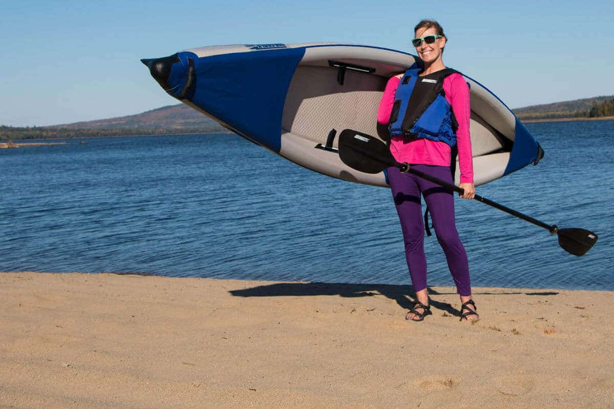 A female kayaker carrying an inflated 393rl RazorLite along a lake shoreline to the put-in.