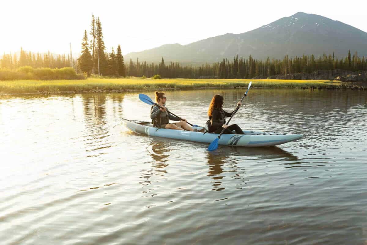 An Aquaglide Cirrus Ultralight 150 tandem inflatable kayak being paddled by a couple on a calm lake at sunset.